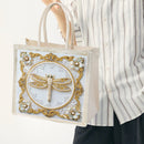 A person in a white and gray striped shirt holds a clear plastic tote bag with a decorative front panel. The bag has beige handles, a white lace background, gold ornate borders, and a beaded dragonfly design in the center.