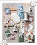 A styled flat-lay of vintage-inspired stationery and decorative paper goods on a textured gray background, including layered envelopes, cards, tags with butterfly and floral cutouts, small white figurines, a column ornament, and paper flowers.