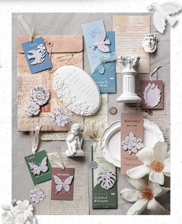 A styled flat-lay of vintage-inspired stationery and decorative paper goods on a textured gray background, including layered envelopes, cards, tags with butterfly and floral cutouts, small white figurines, a column ornament, and paper flowers.