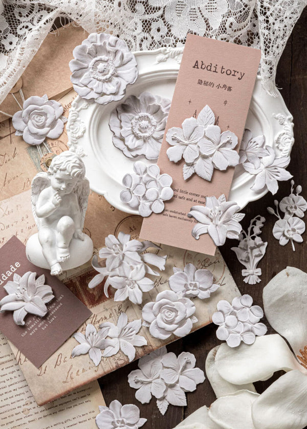 White floral nail art stickers arranged on a vintage-style book, white ceramic plate, lace fabric, and a small white figurine, with two pink cards displaying the brand 'Abditory'.