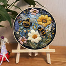 A round embroidered floral and bee artwork displayed in a wooden hoop on a small stand, set on a wooden surface with a plant leaf and a decorative figurine nearby.