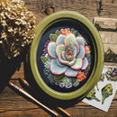 An oval framed floral embroidery or needlework piece featuring a large pale blue-green succulent flower with pink and orange outlines, surrounded by green and purple leaves, displayed on a rustic wooden surface with dried flowers, paintbrushes, and printed botanical cards.