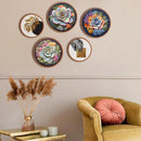 Five circular framed wall art prints with colorful floral and botanical designs are arranged on a beige wall above a mustard yellow armchair with a pink cushion, next to a side table with dried pampas grass.