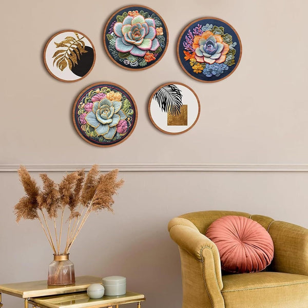 Five circular framed wall art prints with colorful floral and botanical designs are arranged on a beige wall above a mustard yellow armchair with a pink cushion, next to a side table with dried pampas grass.