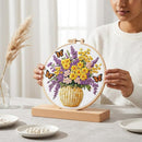 A person in a white sweater holds a round embroidery hoop displaying a colorful floral bouquet with yellow and purple flowers, green leaves, and orange butterflies in a woven basket pattern, mounted on a wooden stand on a table.