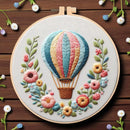 A circular embroidery hoop displays a colorful hot air balloon surrounded by embroidered flowers on a light fabric background, set against a dark brown wooden surface with small scattered flowers around it.
