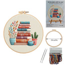Embroidery DIY art kit shown with a completed circular embroidery hoop featuring stacked books, potted plants, and a teacup on a cream fabric background. The image also includes a product box labeled 'Embroidery DIY Art Kit', a smaller hoop with a 20cm/8in diameter marker, two metal needles, and a thread holder with multicolored embroidery floss.