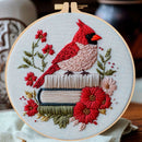 A round wooden embroidery hoop holding a detailed floral and bird embroidery. The design features a red cardinal perched on stacked books, surrounded by red and pink flowers with green leaves on a light beige fabric background.