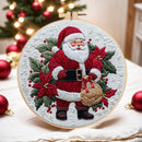 A round embroidery hoop featuring a festive Christmas design of Santa Claus surrounded by green holly leaves, red poinsettias, and white snowflakes on white fabric. The hoop is displayed on a table with blurred red Christmas ornaments and warm lights in the background.