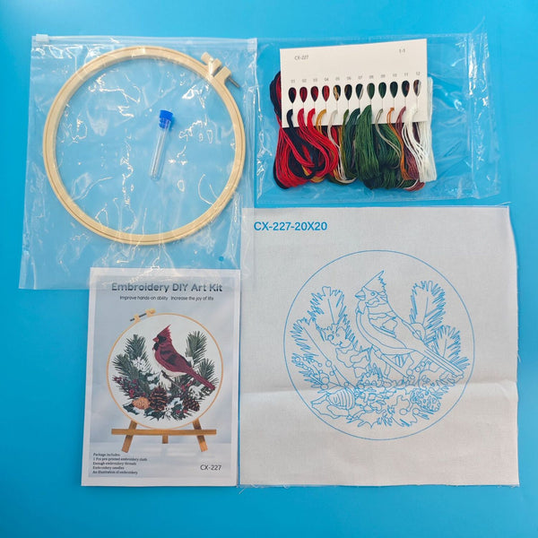 Embroidery DIY art kit laid out on a bright blue background, including an embroidery hoop with thread, a package of multicolor embroidery floss, a pattern booklet showing a cardinal design, and a white fabric square with a blue cardinal embroidery outline.