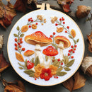 A circular embroidery hoop displays a detailed floral and mushroom embroidery design on white fabric, surrounded by autumn leaves and acorns on a dark gray wooden surface.