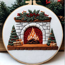 A circular embroidery hoop featuring a festive Christmas fireplace design with white stitched brick, a red and orange fire, green garland with red berries, two decorated trees, and wrapped gifts, shown against a holiday background.