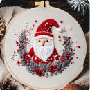 A round embroidery hoop displays a festive Christmas-themed stitched design featuring a Santa Claus figure with a red hat and white beard, surrounded by a gray wreath with red berries, on a cream fabric background.
