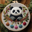 A round embroidery hoop featuring a stitched panda bear sitting on a branch with green leaves, red berries, and hanging Christmas ornaments. The hoop is photographed against a festive background with pine branches and warm wooden elements.