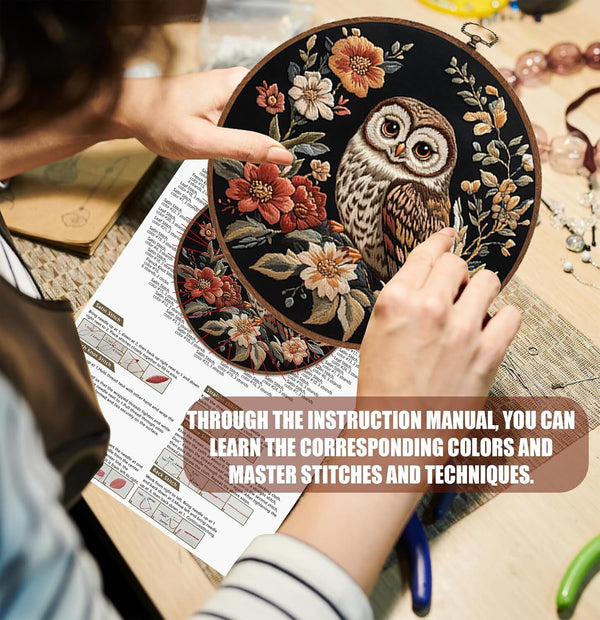 Person holding a round embroidered piece with an owl design, surrounded by crafting materials and an instruction manual.