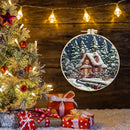 A circular embroidered Christmas ornament depicting a snowy cabin in a pine forest, displayed next to a decorated Christmas tree and wrapped gifts against a rustic wooden wall with warm string lights.