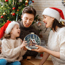 A festive family portrait featuring a father, mother, and child wearing Santa hats, gathered in front of a decorated Christmas tree. The mother holds up a round embroidered Christmas ornament depicting a snowy cabin under a starry night sky, which the father and child are looking at with joyful expressions.