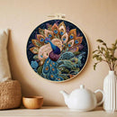 A circular embroidered wall hanging featuring a peacock with spread feathers in rich blue, teal, gold, and purple tones, displayed on a beige wall above a shelf with a white teapot, wooden bowl, vase, and cushion.