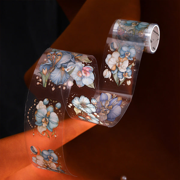 A roll of clear decorative tape with blue, white, and pink floral designs, shown partially unrolled over an orange surface against a dark brown background.