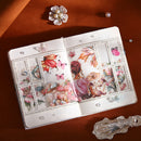 An open decorative journal or notebook spread on a warm brown surface, featuring layered 3D pop-up illustrations of butterflies, flowers, and a woman in pink. The scene is styled with a pearl flower pin, a pearl bead, and a silver spoon around it.