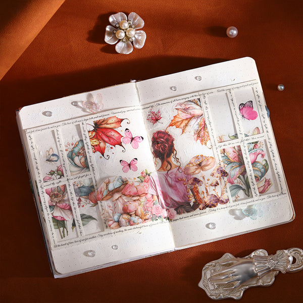 An open decorative journal or notebook spread on a warm brown surface, featuring layered 3D pop-up illustrations of butterflies, flowers, and a woman in pink. The scene is styled with a pearl flower pin, a pearl bead, and a silver spoon around it.
