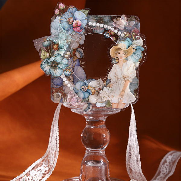 A decorative clear acrylic picture frame with a floral and butterfly embellishment border, a printed illustration of a woman in a white dress, and white pearls, displayed on a clear glass pedestal against a warm brown background.