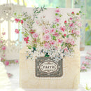 A decorative greeting card or art print featuring a cream background with a large arrangement of pink and white flowers and green vines. The lower portion shows a textured cream envelope or card base with a central gray label that reads “FAITH” in an ornate oval frame with pearl-like accents.