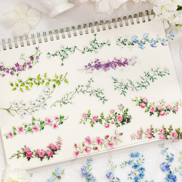 A spiral-bound notebook page decorated with delicate watercolor floral garlands in pink, purple, blue, and green, styled with soft white flowers around it.