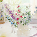 A decorative floral arrangement made from layered translucent paper cutouts shaped like different flower sprigs, displayed in a clear glass vase on a light tabletop.