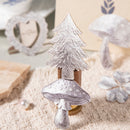 A decorative Christmas tree and mushroom candle holder with a textured silver finish, displayed on a beige woven fabric surface with rustic props in the background.