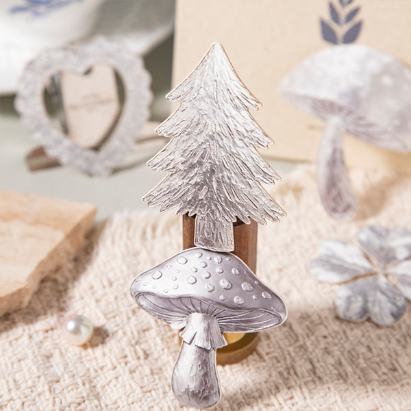 A decorative Christmas tree and mushroom candle holder with a textured silver finish, displayed on a beige woven fabric surface with rustic props in the background.