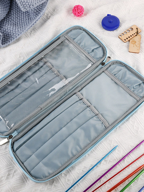 An open light blue and gray knitting needle case with multiple compartments, shown on a white textured blanket with knitting accessories like colorful needles, a blue cable, and wooden tools.