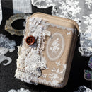 A decorative handmade jewelry box or clutch in a beige linen-like material, embellished with white lace, ribbon bows, faux pearls, a brown button, and floral lace appliques, photographed on a dark surface with lace props around it.