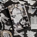 A styled flat-lay of gothic-themed craft embellishments and a clear sticker sheet featuring vintage architectural designs, lace patterns, butterflies, roses, and gothic typography on a dark textured background.