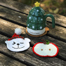 A festive crocheted Christmas-themed set displayed on a weathered wooden table. The set includes a green crocheted mug shaped like a Christmas tree with a yellow star on top, a white crocheted bear face wearing a red and green knit Santa hat, and a white crocheted apple with a red border and green stem.