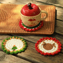 Three handmade crocheted coasters displayed on a rustic wooden table, with a cream-colored ceramic mug featuring a red apple design resting on one of the coasters. The coasters feature autumn-themed designs including a green tree and a brown acorn.