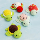 Six crocheted turtle toys in assorted pastel and bright colors are arranged on a light blue surface. The turtles feature green heads and shells with shells in yellow, red, pink, brown, and white, each with small black bead eyes.