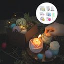 A set of flower-shaped wax candles in soft pastel colors, styled in a dark setting with a hand lighting one with a match. A brown box containing wrapped candles and decorative greenery sits beside the lit candles, and a circular inset shows additional wrapped candle colors and accessories.