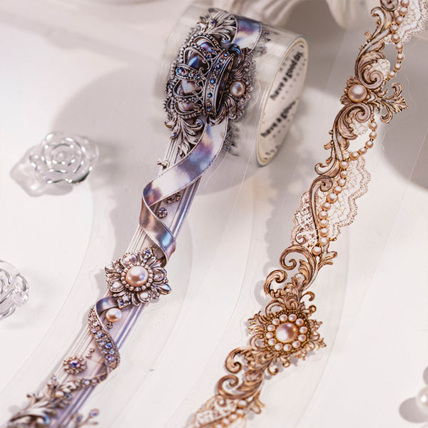 Two decorative patterned rolls of washi tape displayed on a clean white background. One roll features an iridescent silver and blue filigree design with faux pearls, and the other shows a warm gold-toned lace and scroll pattern.