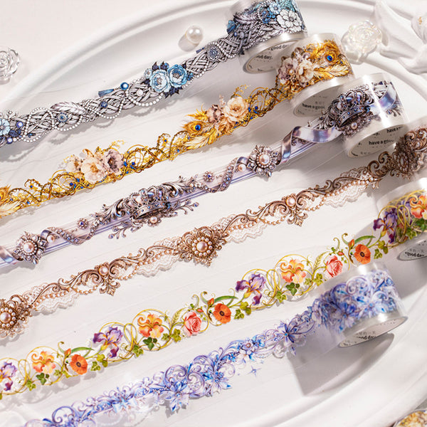 A close-up of multiple rolls of decorative washi tape laid out on a white surface. The tapes feature ornate lace-like patterns and floral motifs in silver, gold, bronze, blue, purple, and multicolor designs.