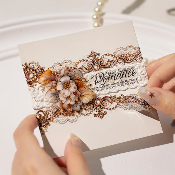 A close-up of a hand holding a decorative card with ornate lace, 3D floral embellishments, and the word 'Romance' printed on it. Another hand is visible at the right edge against a soft white background.