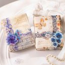 Two decorative gift boxes wrapped in vintage-style patterned paper, placed on a white surface with a pearl necklace and small clear embellishments nearby. The wrapping features floral designs, lace textures, butterflies, and printed text.