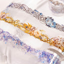 Three decorative lace or trim strips with ornate floral and filigree patterns, shown close up on a white fabric background. The top strip is silver and blue, the middle is gold and peach, and the bottom is purple and blue.