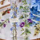 A decorative clear glass vase filled with illustrated floral stems and small paper labels reading words like "journey," "adventure," "explore," and "wanderlust," set against a softly blurred blue floral background.