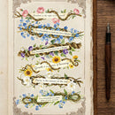 An open vintage-style book or journal page with decorative floral garlands and inspirational quotes, shown beside a wooden surface and a dark fountain pen.