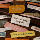 A styled flat-lay of various small, torn fabric or paper labels with inspirational quotes, arranged on a dark wooden background.
