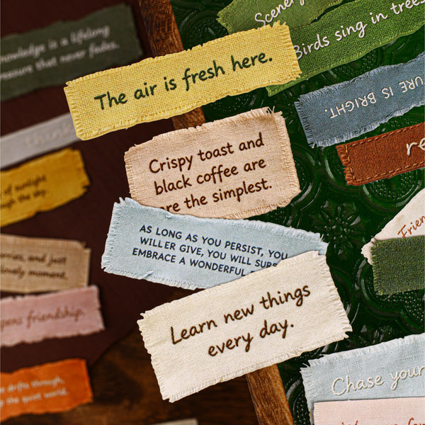 A close-up of multiple small fabric tags with handwritten motivational phrases, arranged on a dark green textured surface.