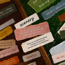 A close-up of a textured dark green leather wall or board decorated with multiple colorful fabric tags and handwritten-style labels with motivational and scenic phrases.