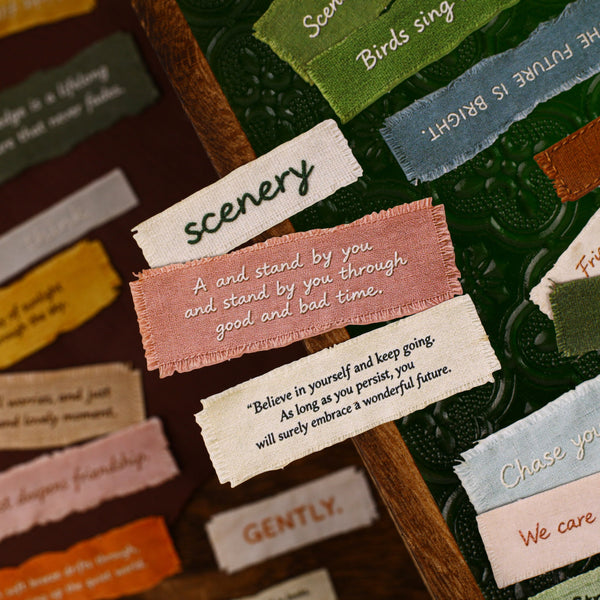 A close-up of a textured dark green leather wall or board decorated with multiple colorful fabric tags and handwritten-style labels with motivational and scenic phrases.