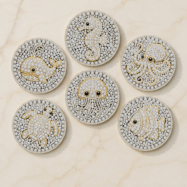 Six round, heavily embellished jewelry pieces arranged on a light marble surface. Each piece features a white base covered in small clear stones, with gold outlines and details forming sea creatures including a seahorse, octopus, crab, fish, and another sea life figure.